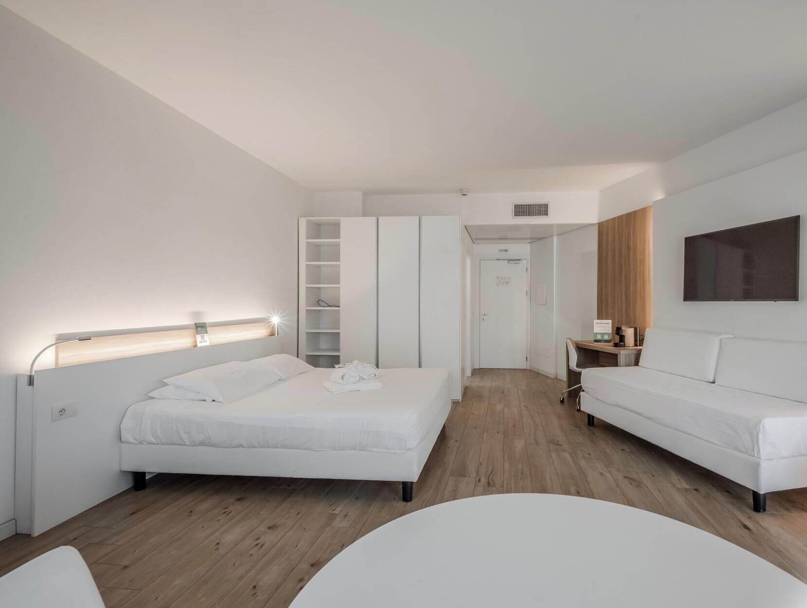 A modern hotel room with a bed, a seating area, a wardrobe, and a television.