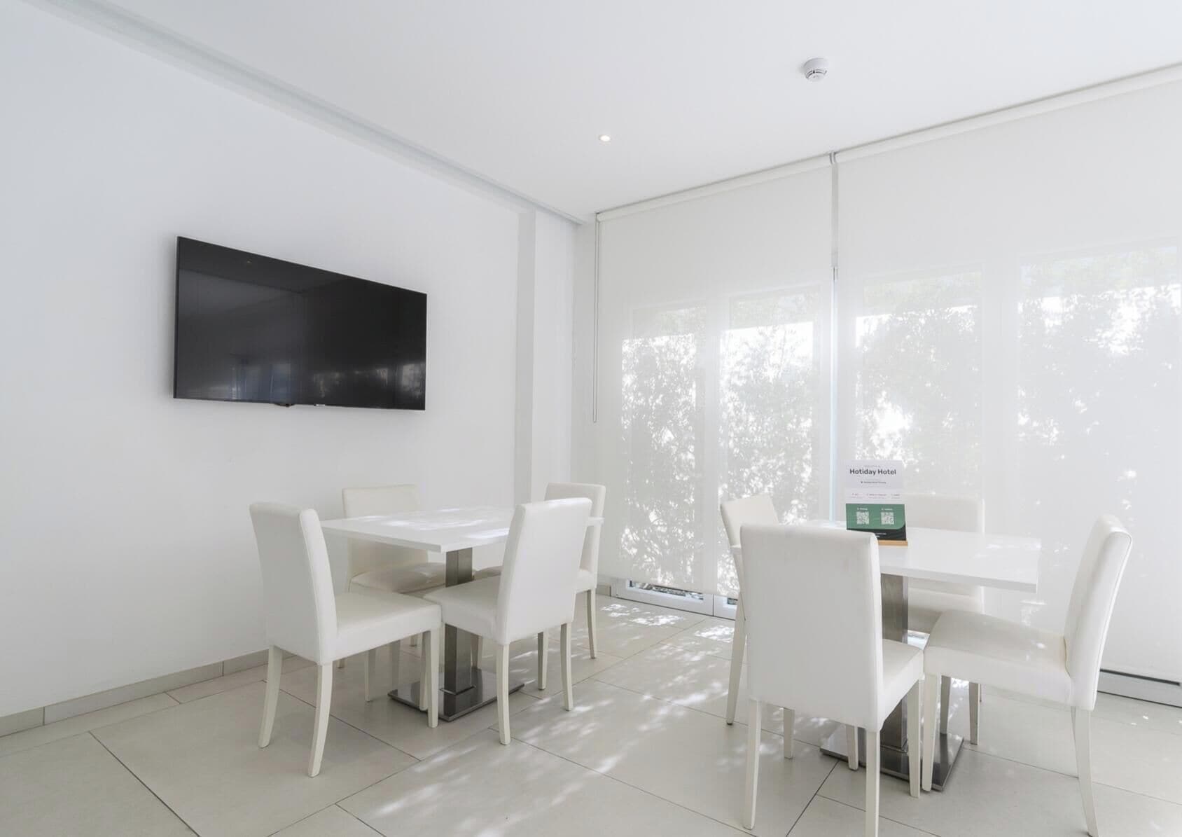 A modern hotel room with a large flat-screen TV mounted on the wall, a dining table with chairs, and a view of trees through the window.