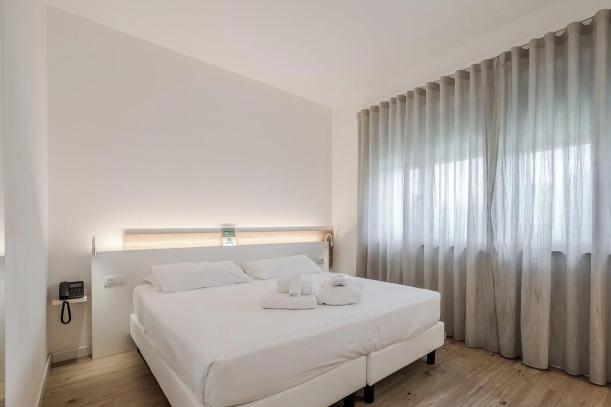 A modern hotel room with a large bed, a bedside table with a phone, and a window with sheer curtains.