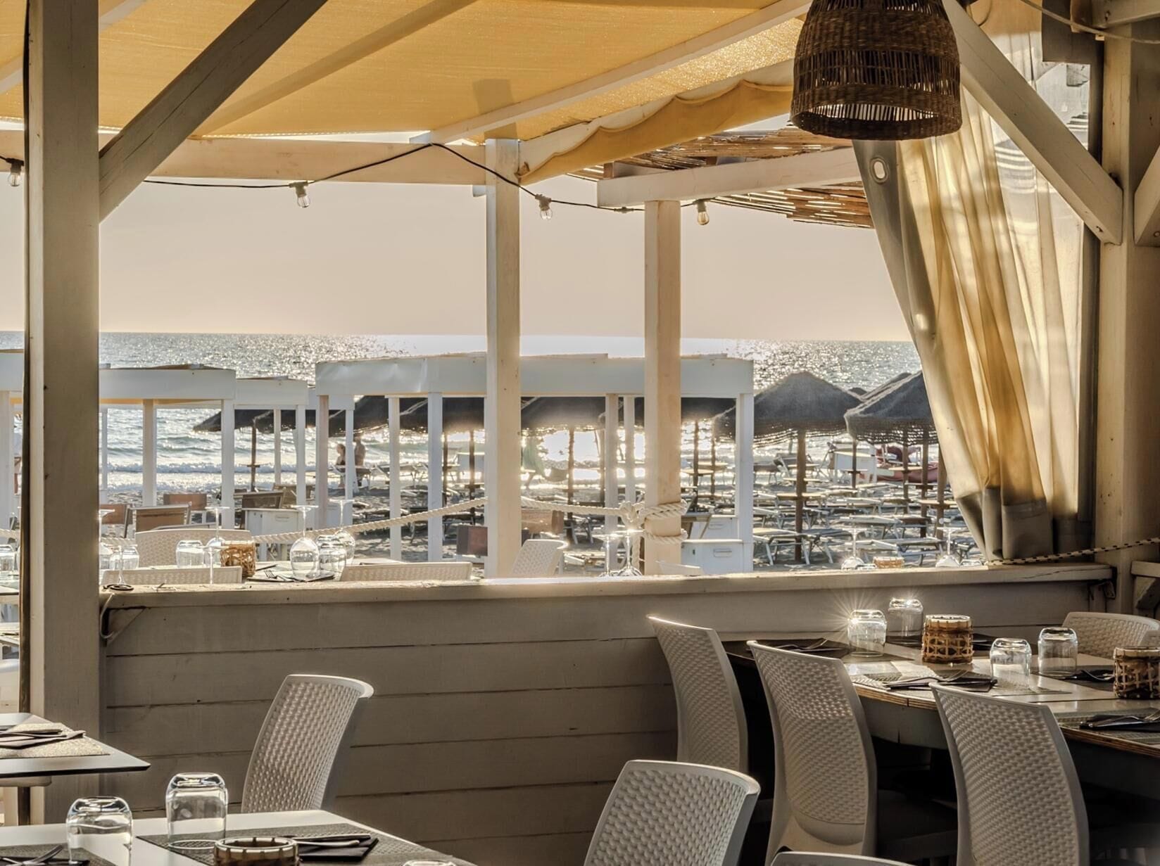 An outdoor dining area with tables and chairs, a view of the beach, and umbrellas.