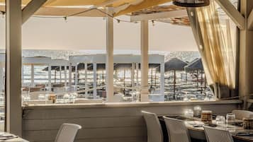 An outdoor dining area with tables and chairs, a view of the beach, and umbrellas.