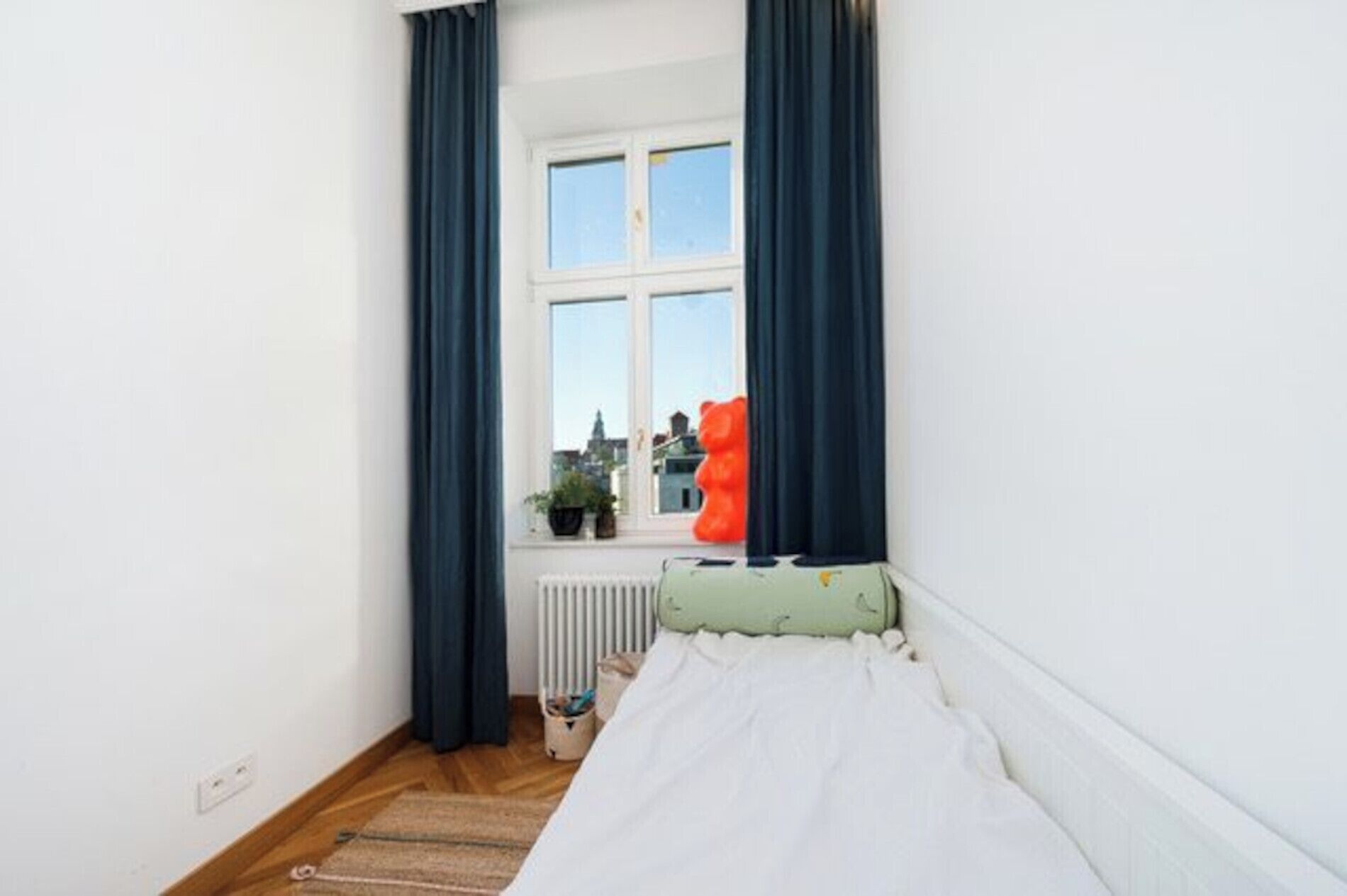 A narrow room with a bed, a radiator, a window with curtains, and a view of buildings outside.