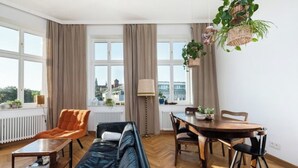 A modern living room with a sofa, a dining table with chairs, a radiator, and large windows with curtains.