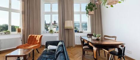 Un salon moderne avec un canapé, une table à manger et des chaises, un radiateur et de grandes fenêtres avec des rideaux.