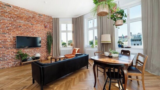 A living room with a brick wall, a flat-screen TV, a black sofa, a wooden dining table with chairs, and hanging plants.