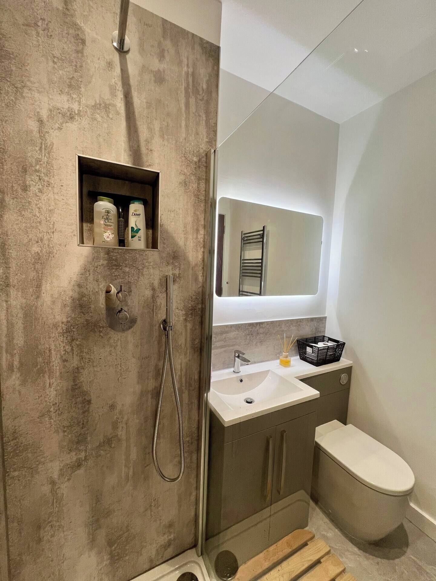 Ein modernes Badezimmer mit einer Duschwand aus Beton, einer eingebauten Ablage für Toilettenartikel, einem Waschtisch mit Spiegel und einer Toilette.