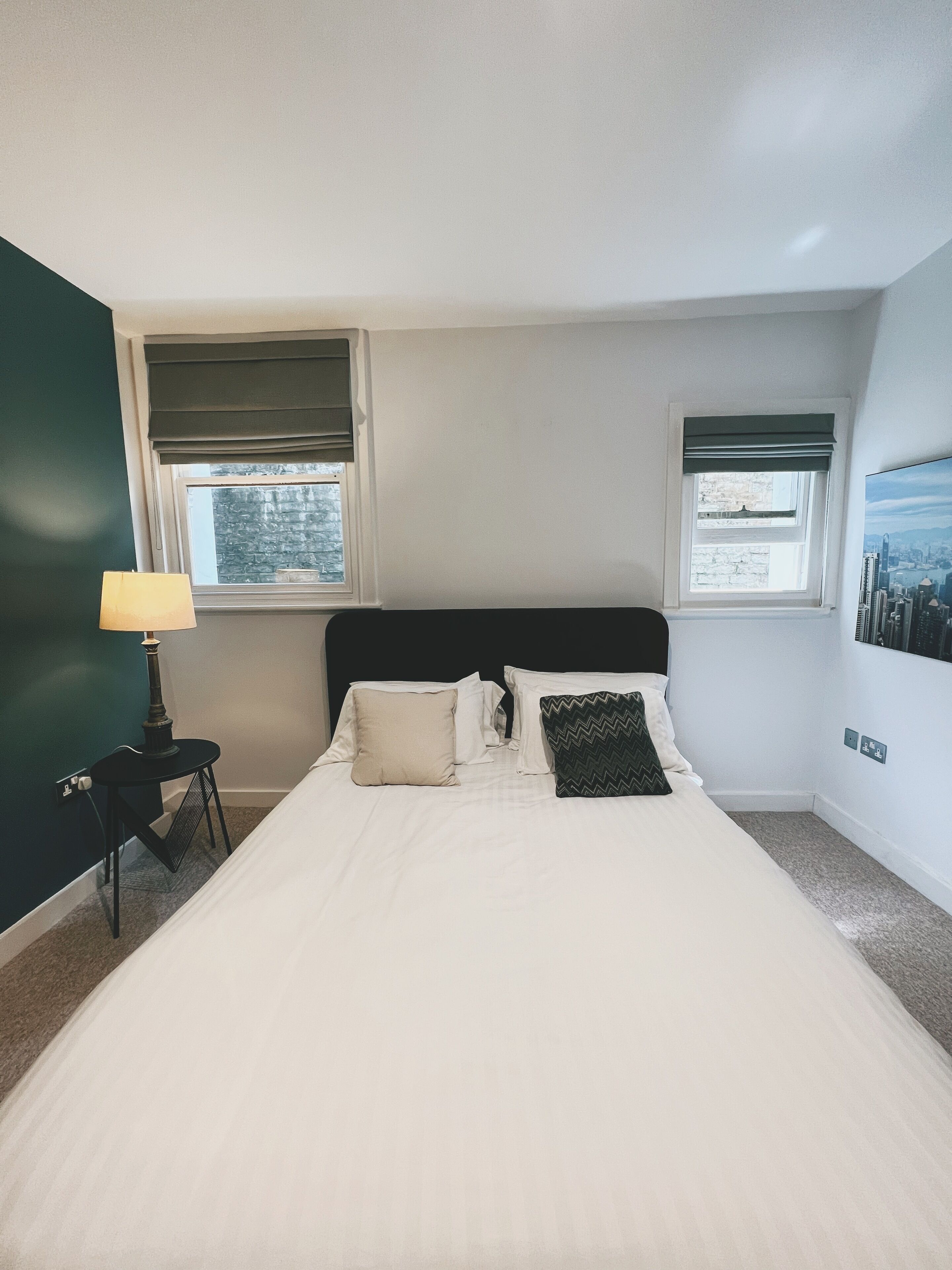A neatly made bed with white bedding and two pillows, a small table with a lamp, and two windows with blinds.