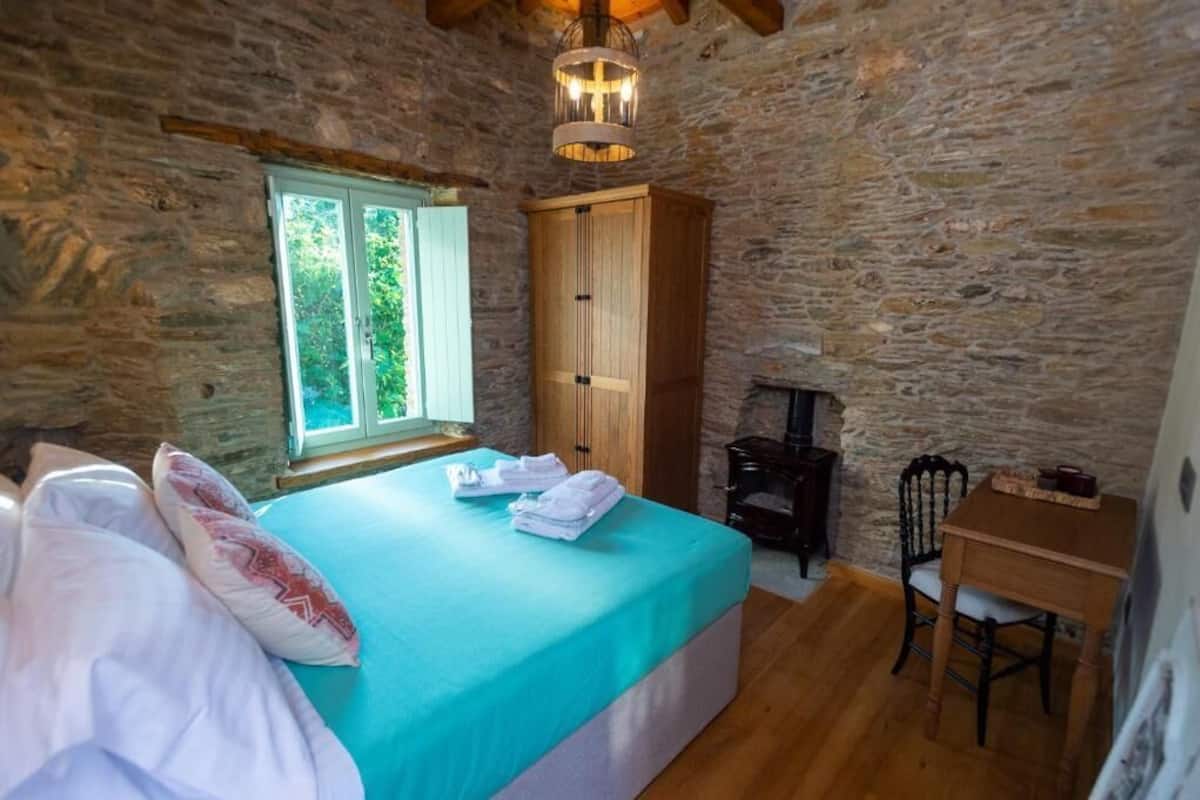 A stone-walled room with a bed, a window, a wooden wardrobe, and a small table with a chair.