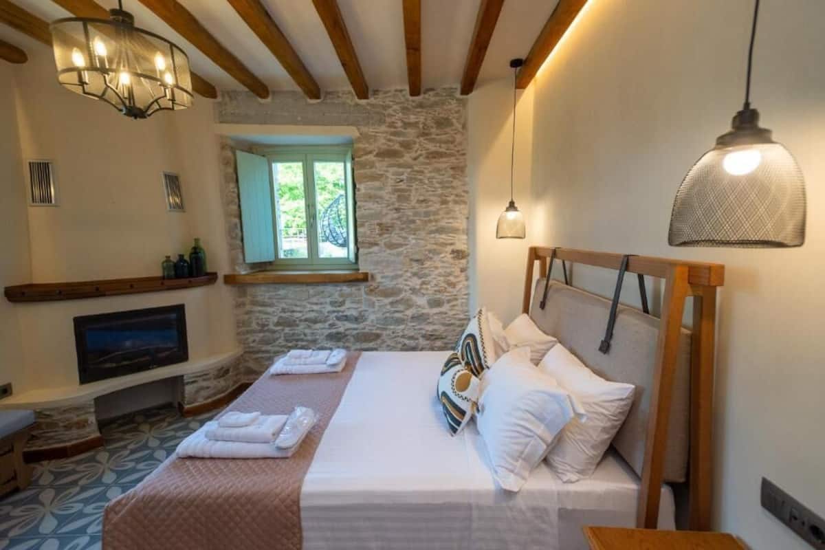 A bedroom with a stone wall, wooden beams, a bed with white bedding, a TV, and a window with a view.