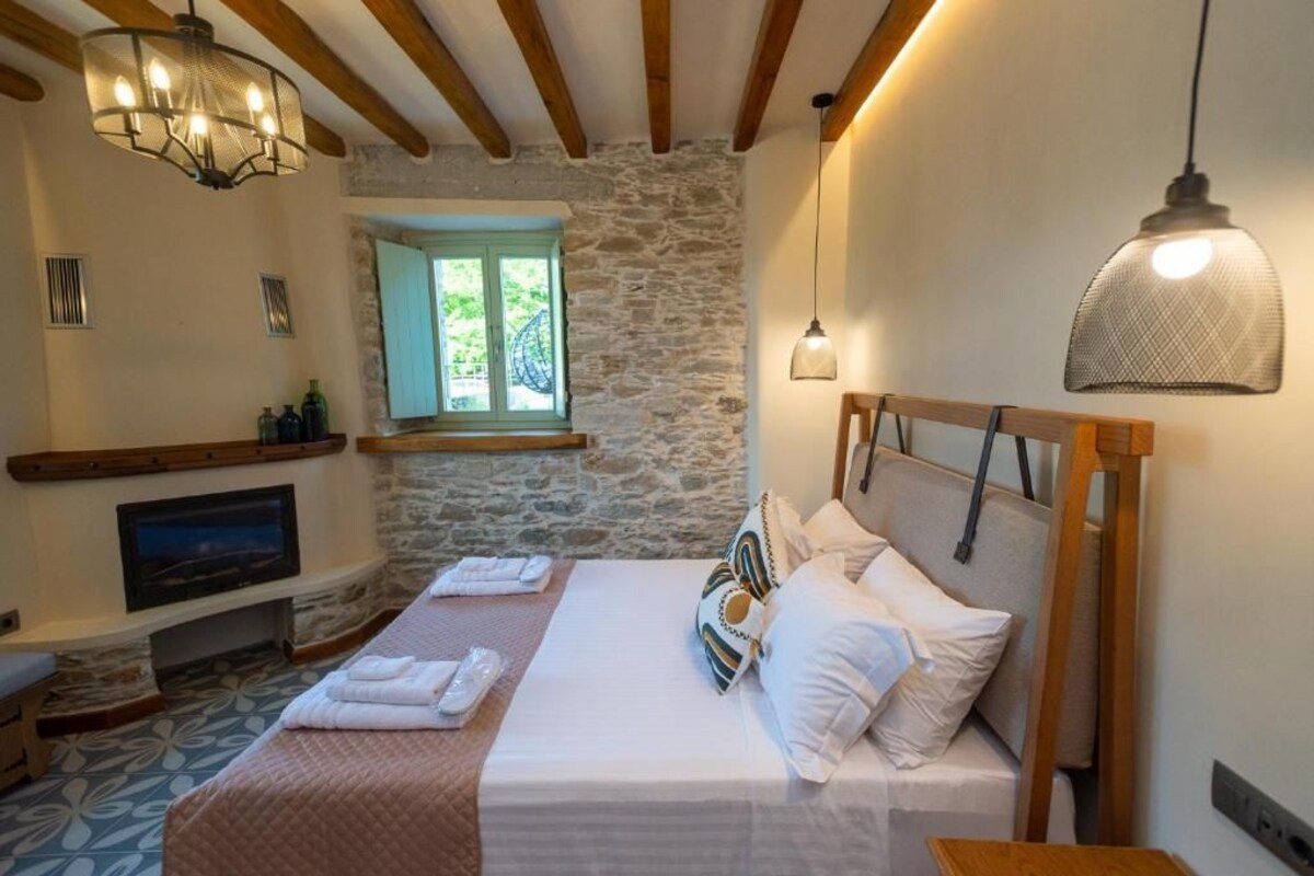 Un dormitorio con pared de piedra, vigas de madera, cama con ropa de cama blanca, un televisor y una ventana con vistas.