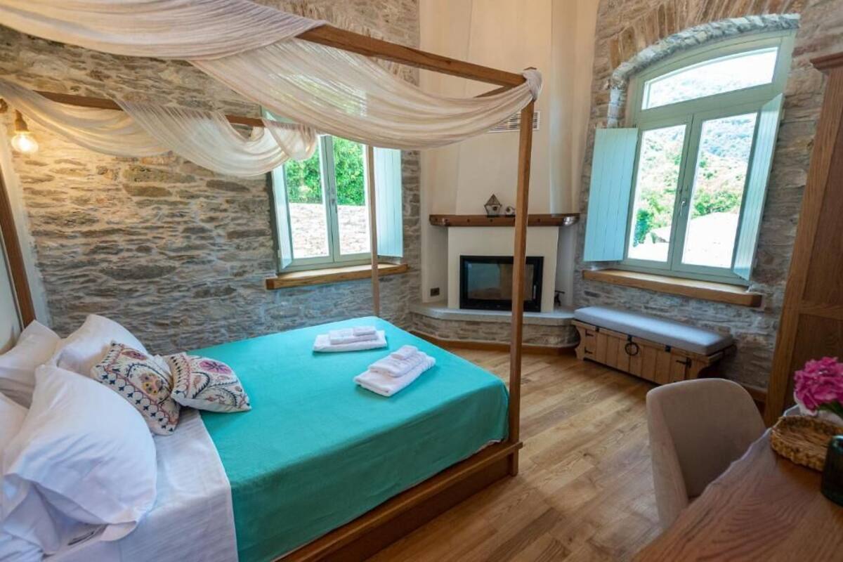 A four-poster bed with a green bedspread, a stone wall, a fireplace, and a bench by a window.