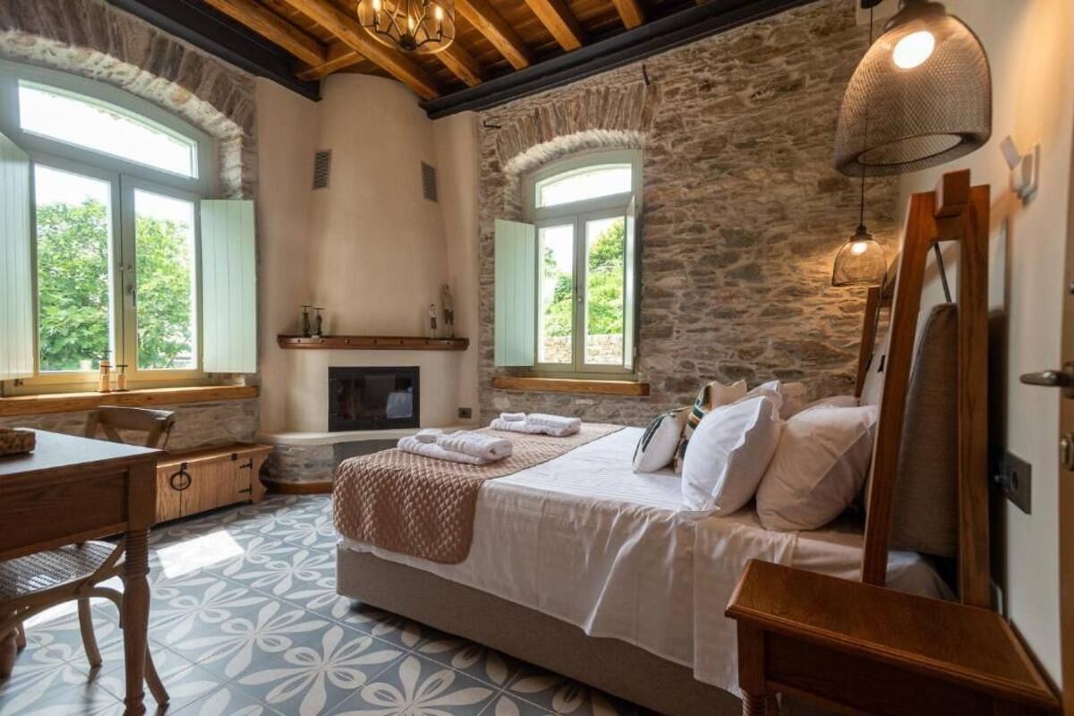 A bedroom with a stone wall, a fireplace, a bed with pillows, a wooden desk, and a chair.