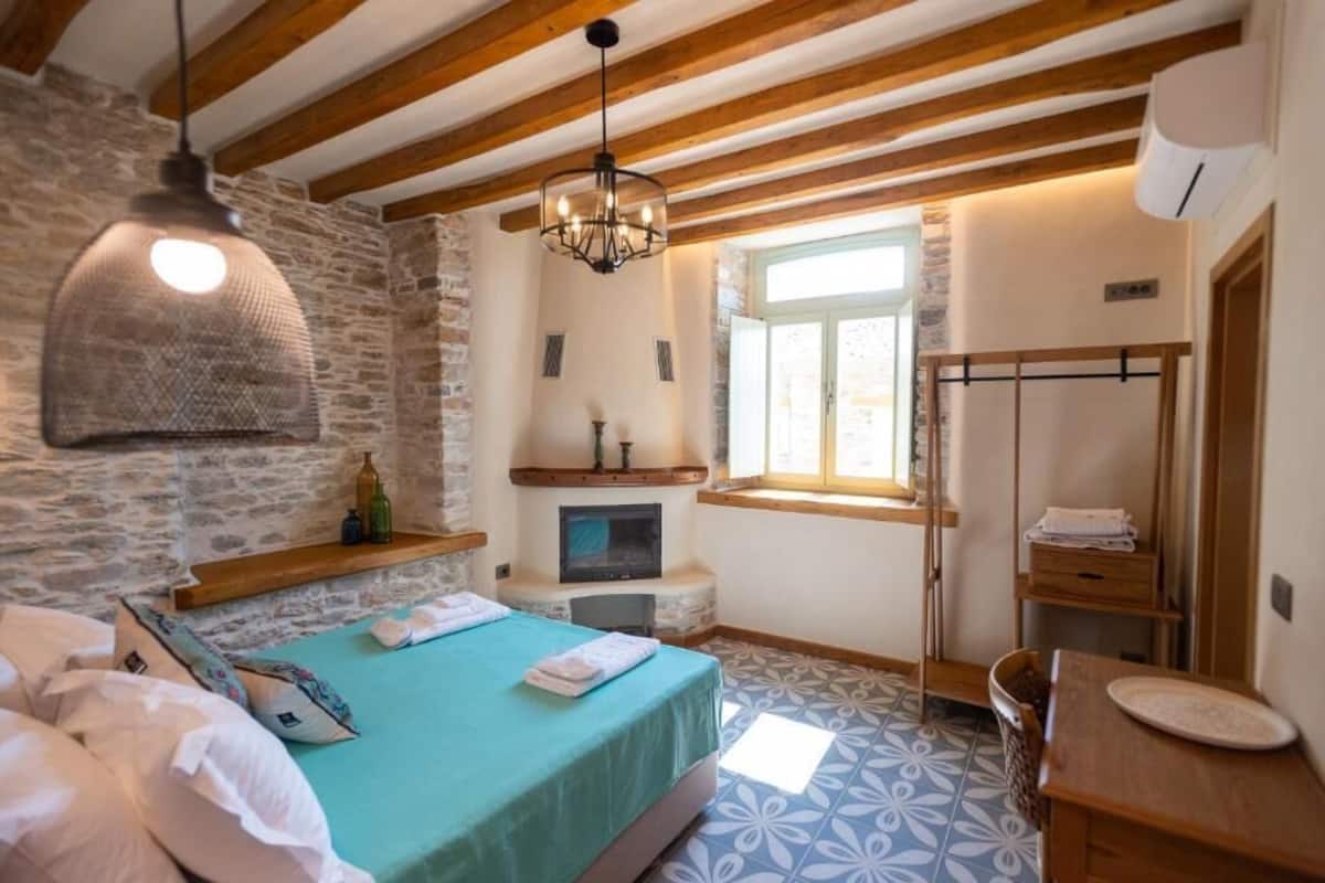 A bedroom with a stone wall, a bed with a green bedspread, a hanging light fixture, a fireplace, and a window.