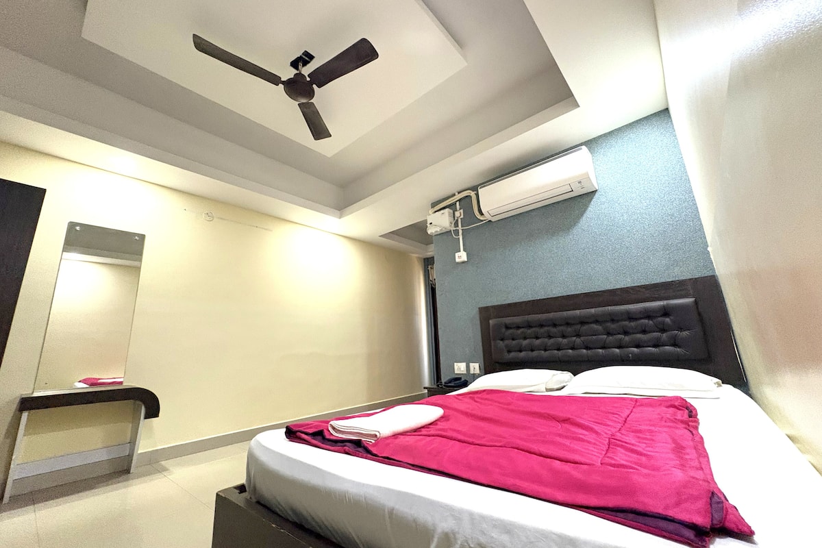 Una habitación de hotel con una cama, ventilador de techo, unidad de aire acondicionado, mesita de noche y tocador.