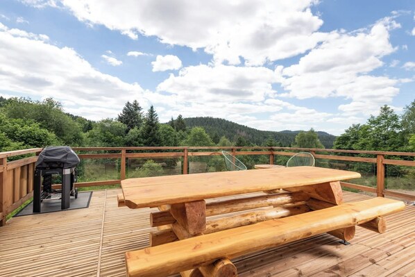 Une terrasse en bois avec une table de pique-nique et un barbecue, entourée d’une rambarde en bois, et offrant une vue sur un paysage boisé.