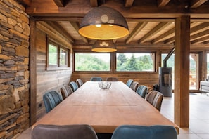 Een lange houten eettafel met stoelen, een stenen muur en een groot raam met uitzicht op groen.