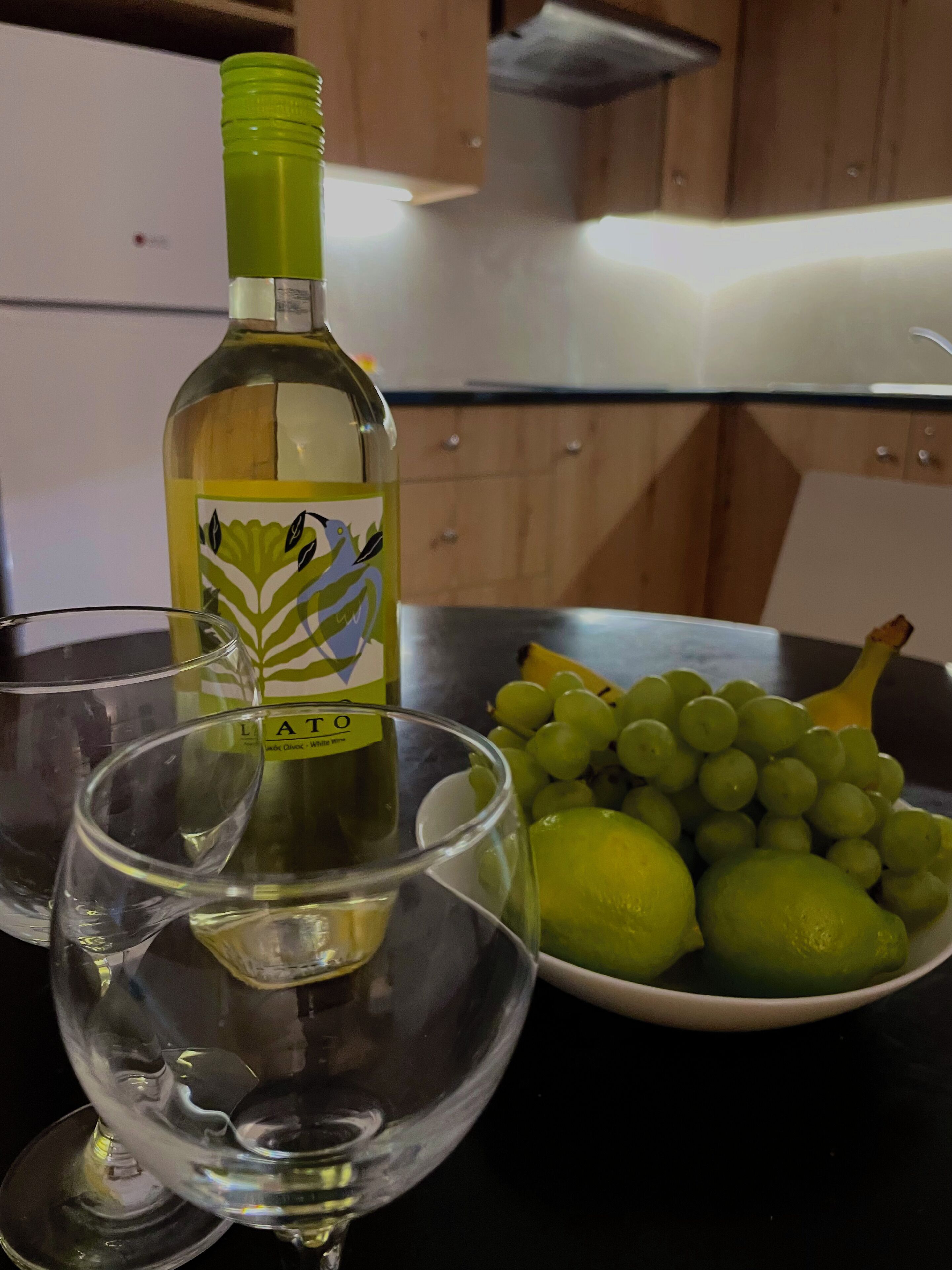 A kitchen counter with a bottle of white wine, a bowl of green grapes, and a glass.
