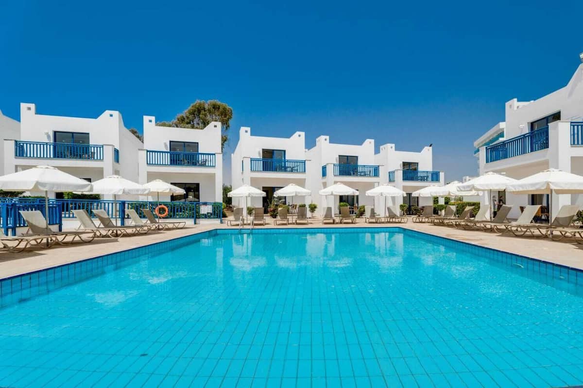 A swimming pool with lounge chairs and umbrellas, surrounded by white buildings with blue railings.
