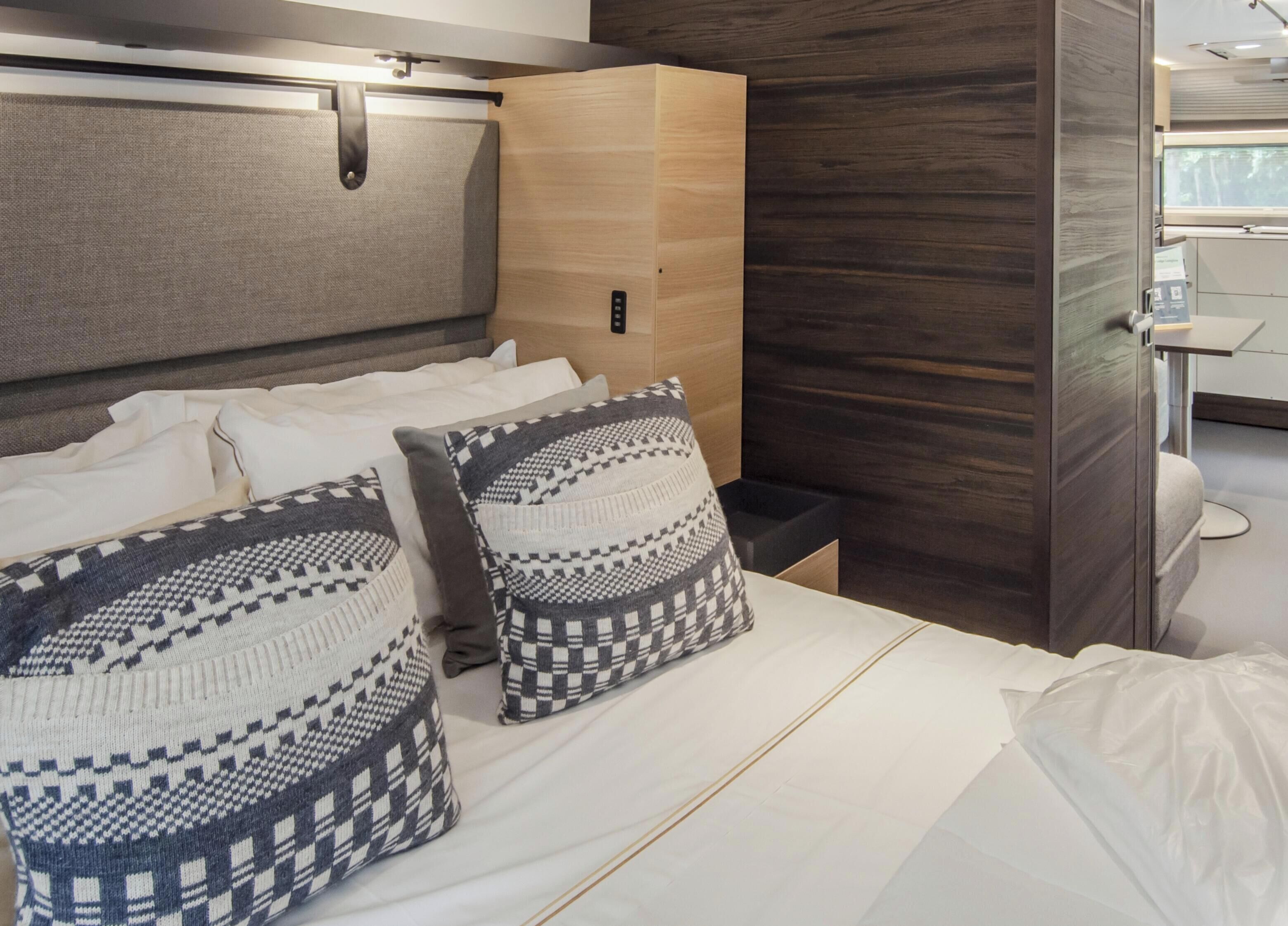 Habitación de hotel moderna con cama, dos almohadas estampadas, una mesita de noche, un armario pequeño y vistas al baño.