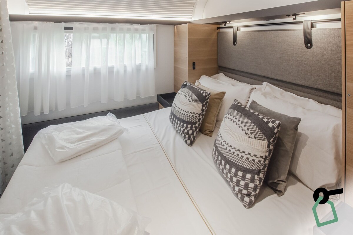 A neatly made bed with patterned pillows, a window with sheer curtains, and a headboard with a light wood finish.