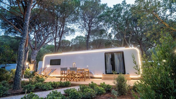 Una motorhome moderna con un porche cubierto, mobiliario de exterior y rodeada de árboles frondosos.