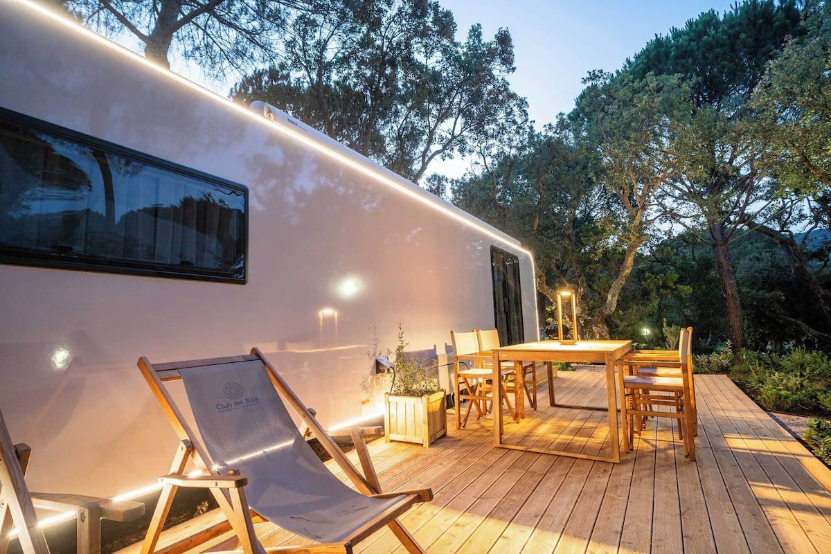 A modern RV with a wooden deck, outdoor seating, and a garden setting.