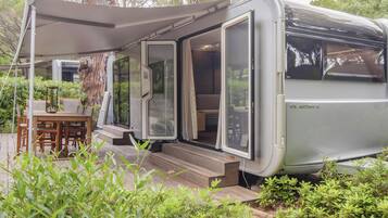 Una autocaravana moderna con un porche cubierto, comedor y un gran árbol de fondo.
