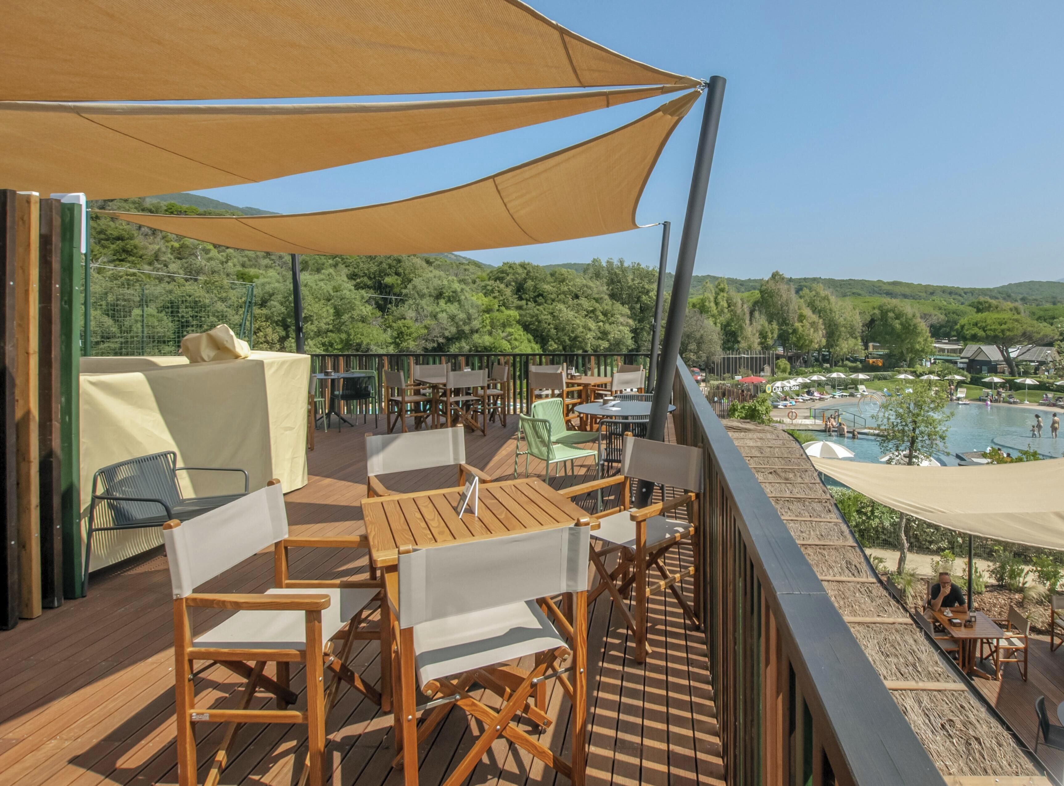 Terraza de madera con mobiliario exterior y vistas a una piscina y colinas.