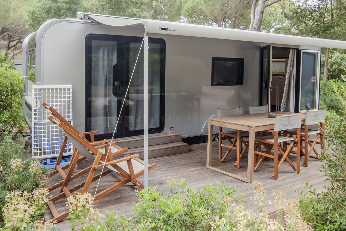 A recreational vehicle with a dining area, including a table and chairs, surrounded by greenery.