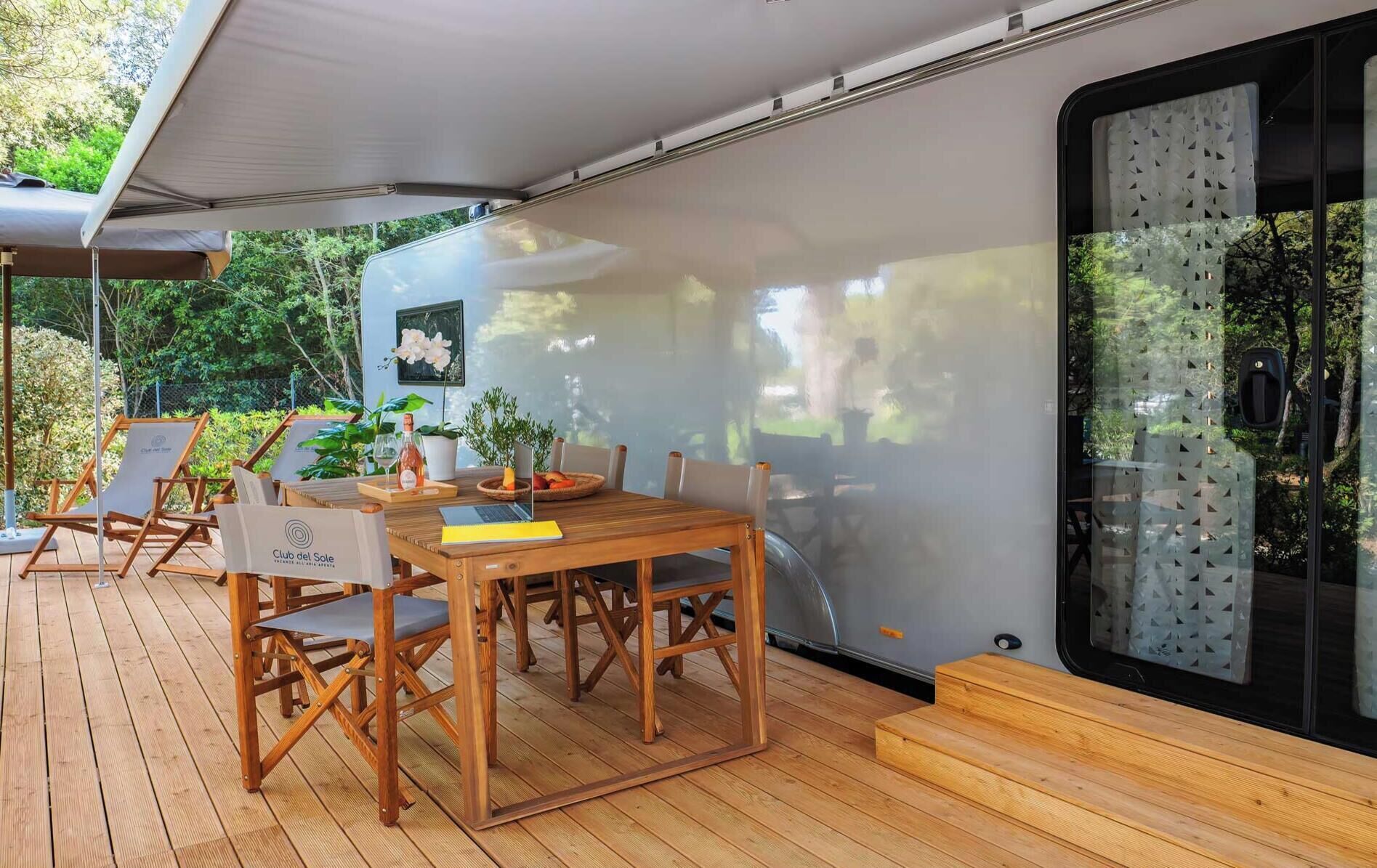 An outdoor dining area with a wooden table and chairs, a laptop, and a bottle of wine, adjacent to a covered seating area with lounge chairs.