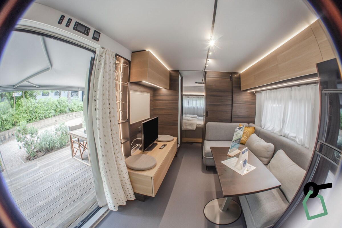 A modern RV interior with a wooden deck, a seating area, a small table, and a TV.