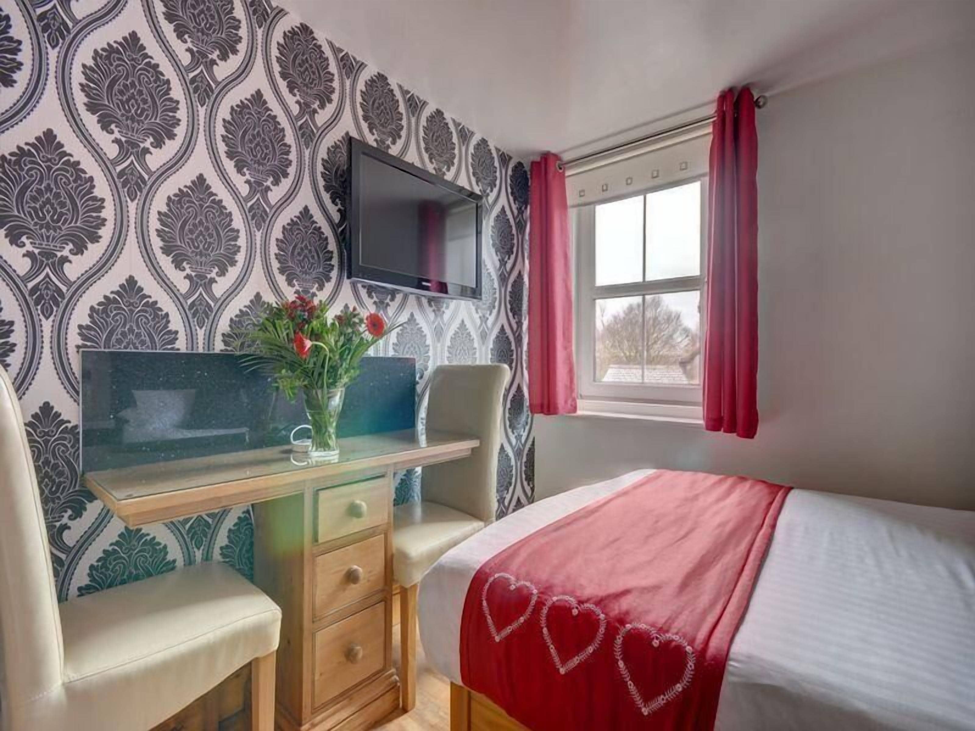 Un dormitorio con cama, escritorio y silla, televisor montado en la pared y ventana con cortinas.