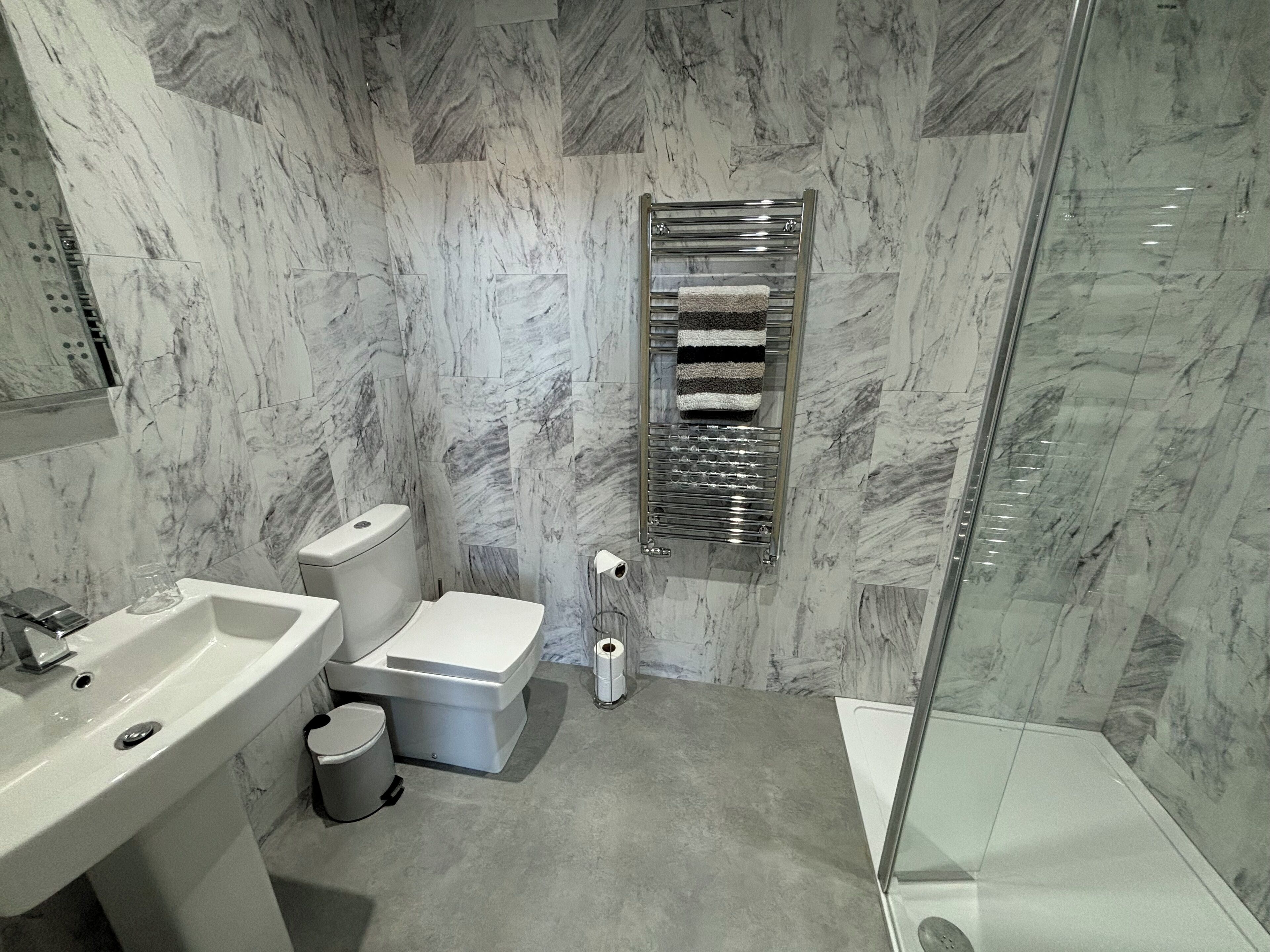 A modern bathroom with a marble wall, a glass shower enclosure, a white sink, a toilet, and a towel rack.