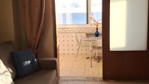 Un salon comprenant un canapé, une table à manger avec des chaises, et une vue sur une terrasse extérieure accessible par une porte coulissante, avec un sol carrelé et une table agrémentée d’un vase de fleurs.
