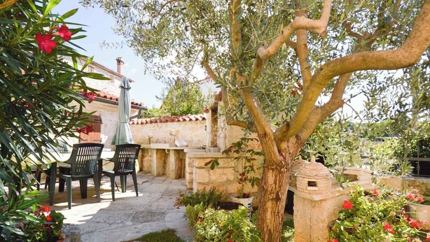 Patio esterno con pavimento in pietra, muro in pietra e un albero con una casetta per uccelli. Si vedono due sedie e un tavolo.