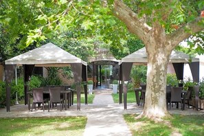 A garden patio with tables and chairs under pergolas, a central pathway, and a large tree.