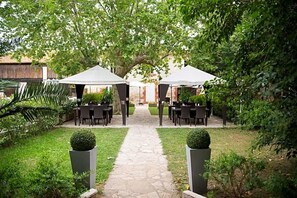 A garden pathway leading to two covered seating areas with tables and chairs, surrounded by greenery and a thatched-roof structure in the background.