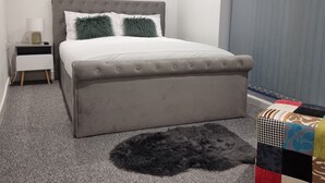 A bedroom with a grey upholstered bed, a small bedside table with a lamp, a grey furry rug, and a colorful ottoman. - London Oasis Modern 2BR Apartment (London)
