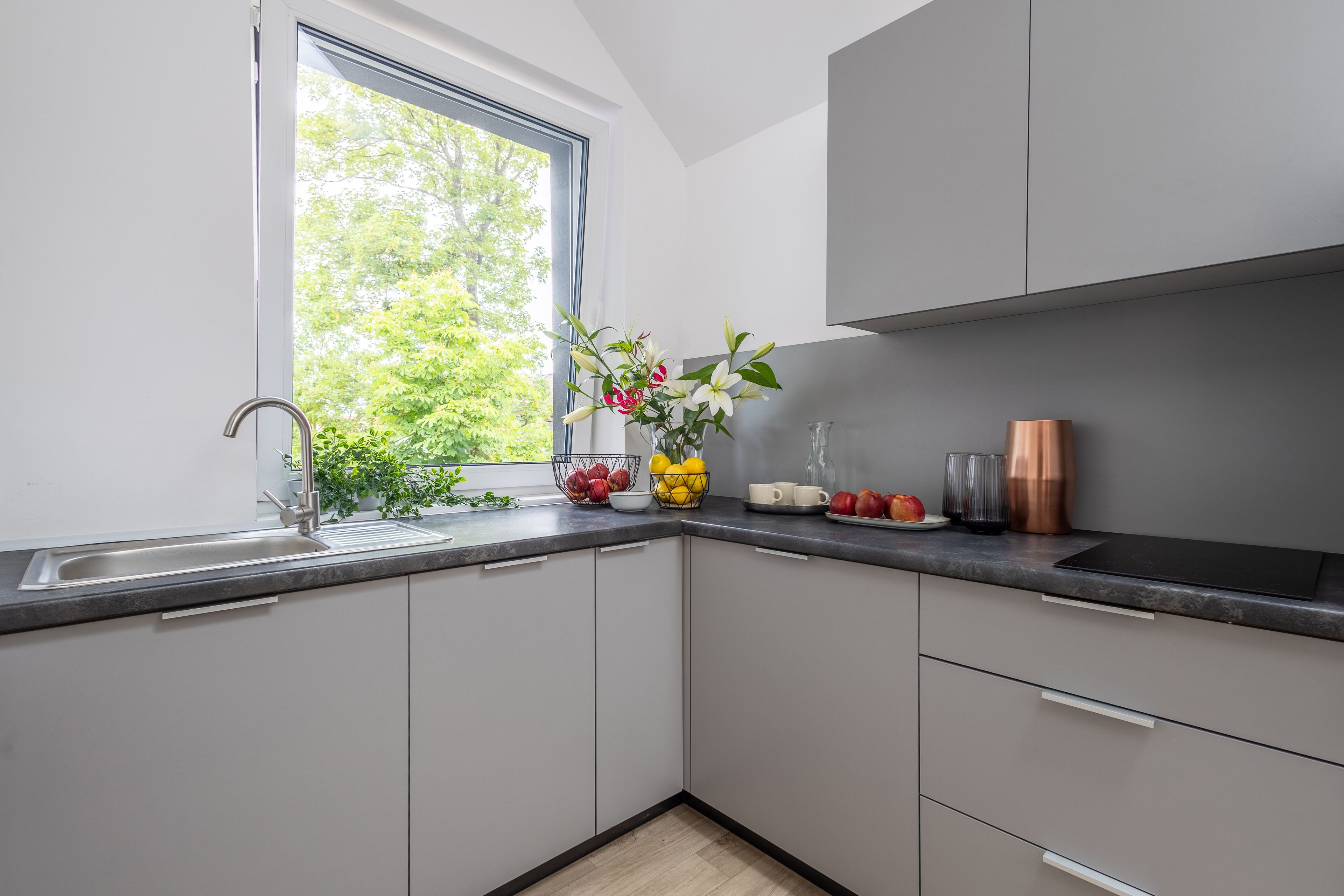 Une cuisine moderne avec une fenêtre, des placards gris, un évier et un plan de travail sur lequel sont disposés divers objets.