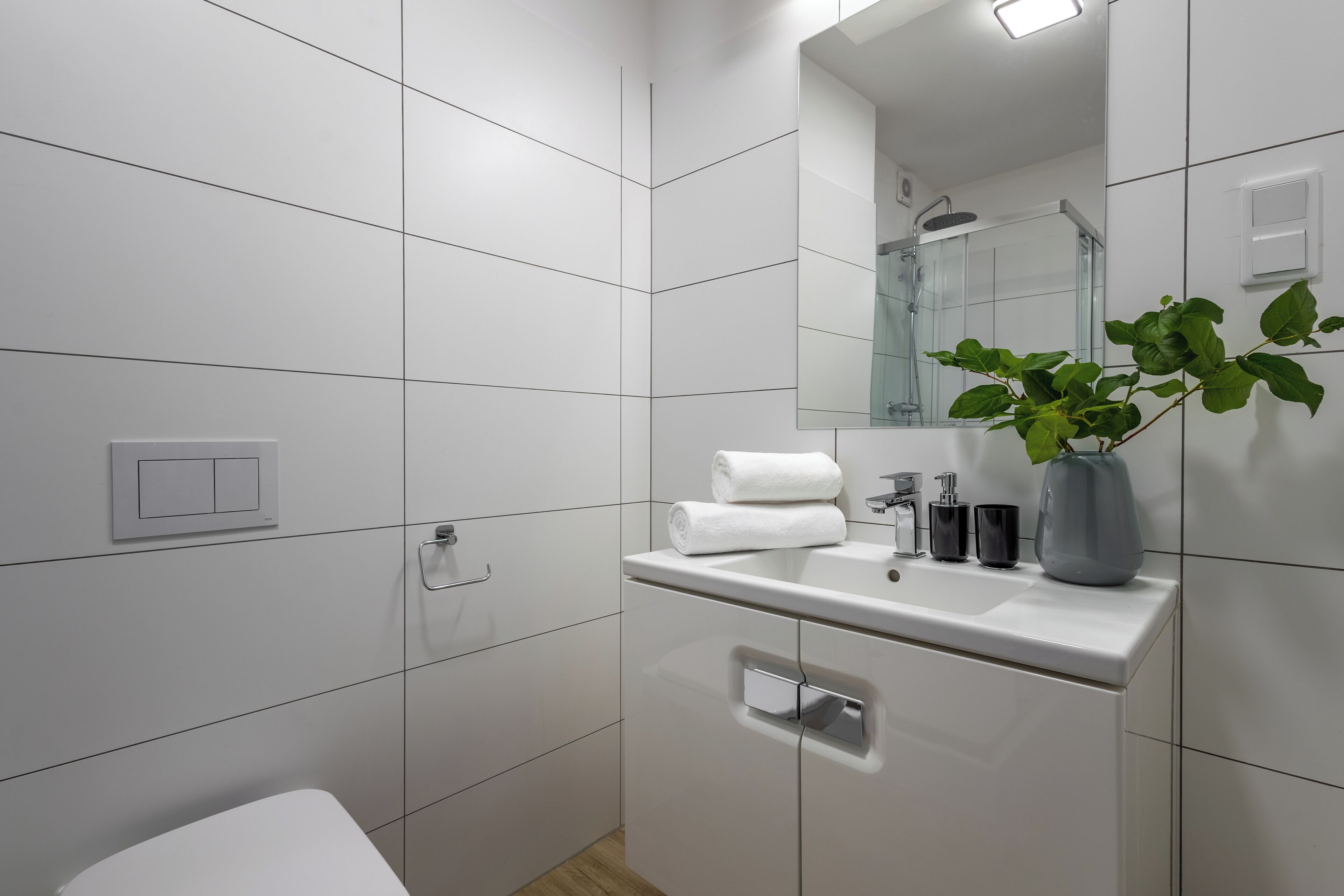 A modern bathroom with white tiles, a white sink, a mirror, a towel, a plant, and a shower.