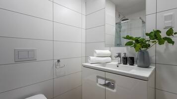A modern bathroom with white tiles, a white sink, a mirror, a towel, a plant, and a shower.
