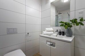 Un baño moderno con azulejos blancos, un lavabo blanco, un espejo, una toalla, una planta y una ducha.