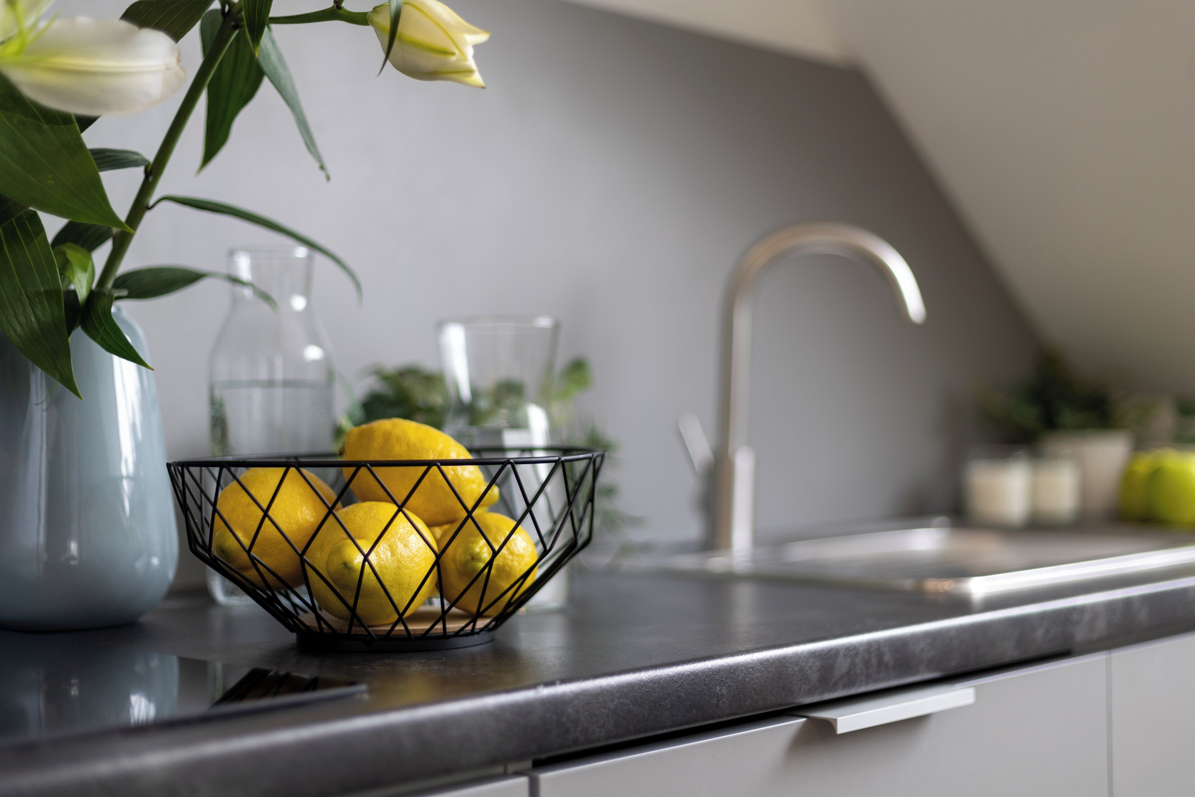 A kitchen counter with a wire basket containing lemons, a vase with white flowers, and a glass pitcher.