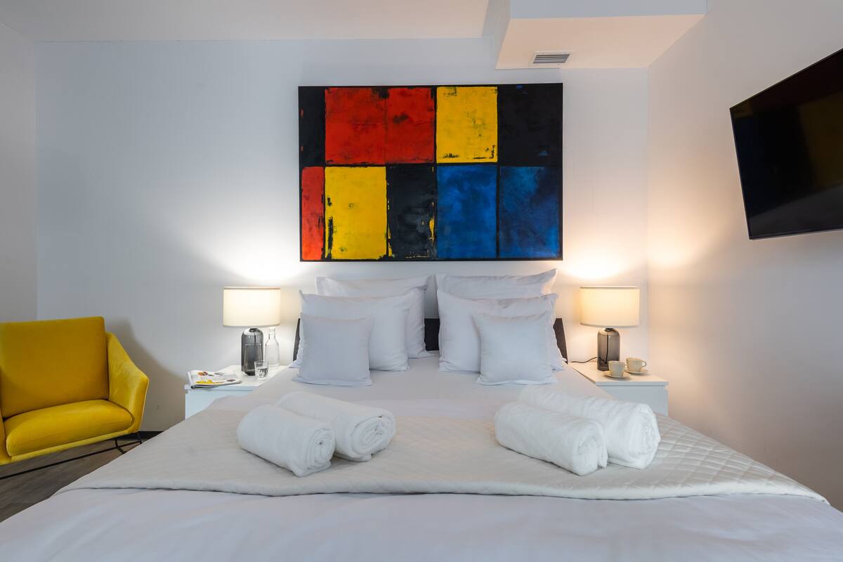 Una habitación de hotel con una cama grande, almohadas blancas, dos mesitas de noche con lámparas, un televisor de pantalla plana y un sillón amarillo.