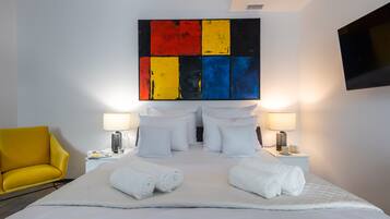 Una habitación de hotel con una cama grande, almohadas blancas, dos mesitas de noche con lámparas, un televisor de pantalla plana y un sillón amarillo.