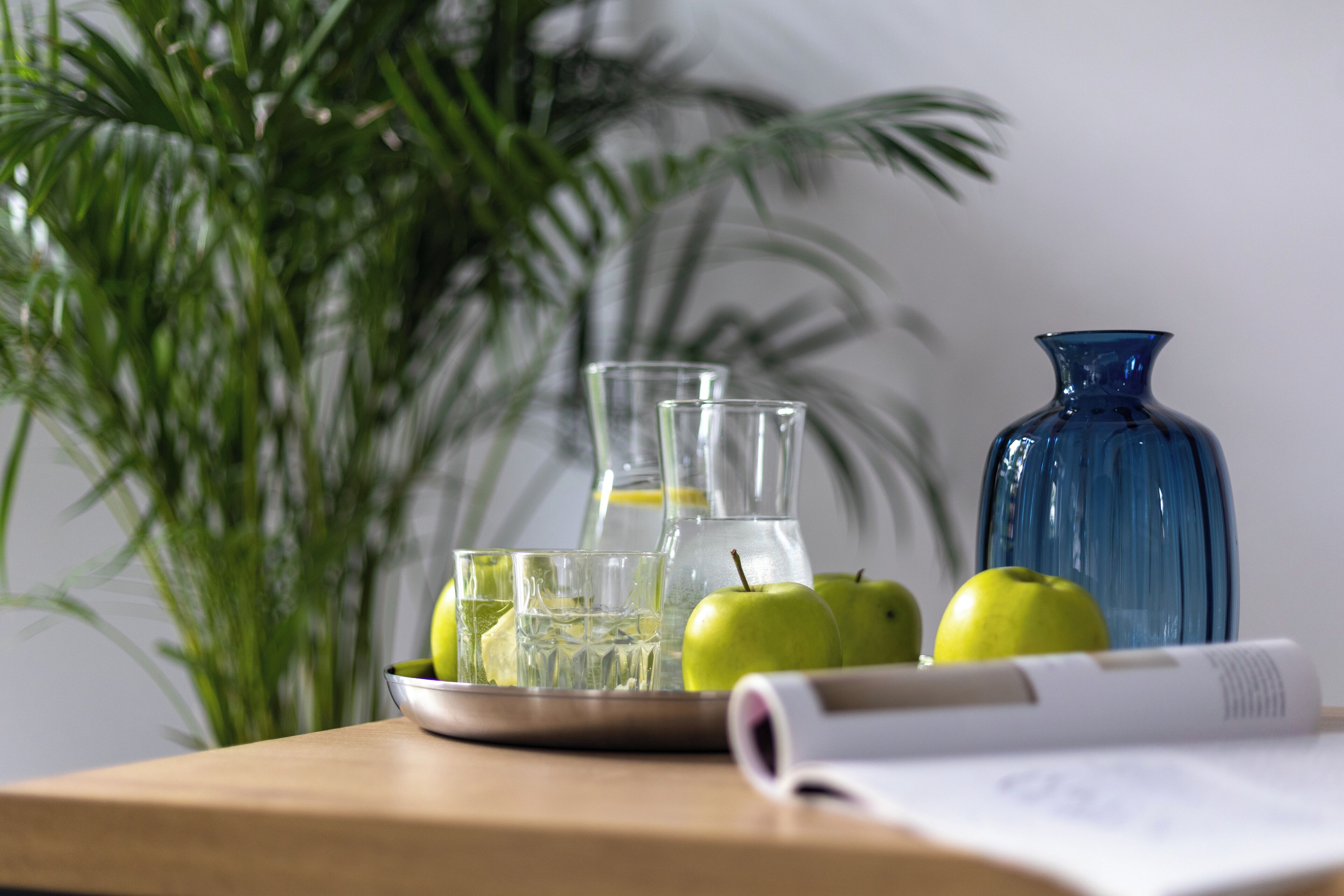 A table with a plate of green apples, a glass pitcher, and a blue vase.