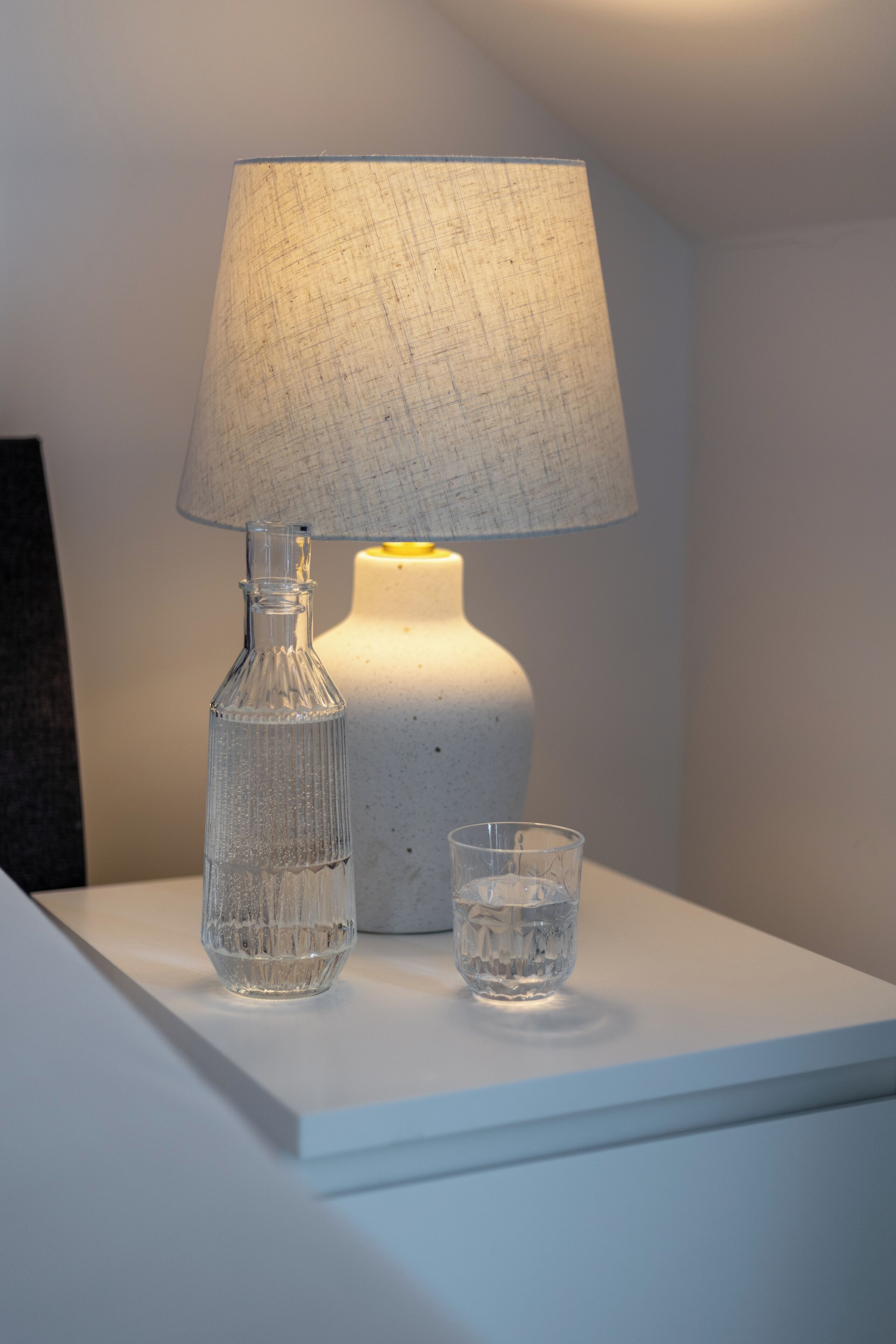 A white table with a clear glass bottle, a glass, and a lamp with a white shade.
