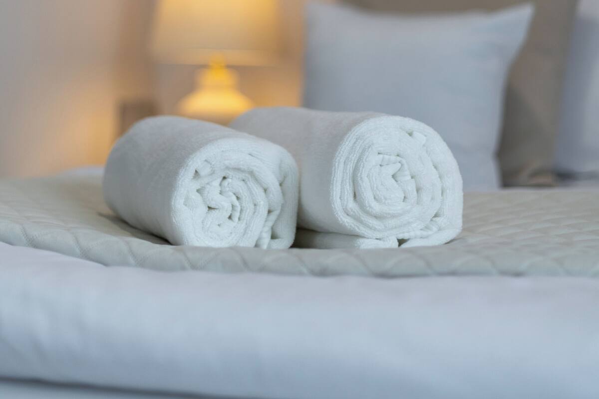 Una cama bien hecha con toallas blancas enrolladas, una lámpara de noche y una pared con cuadros enmarcados.