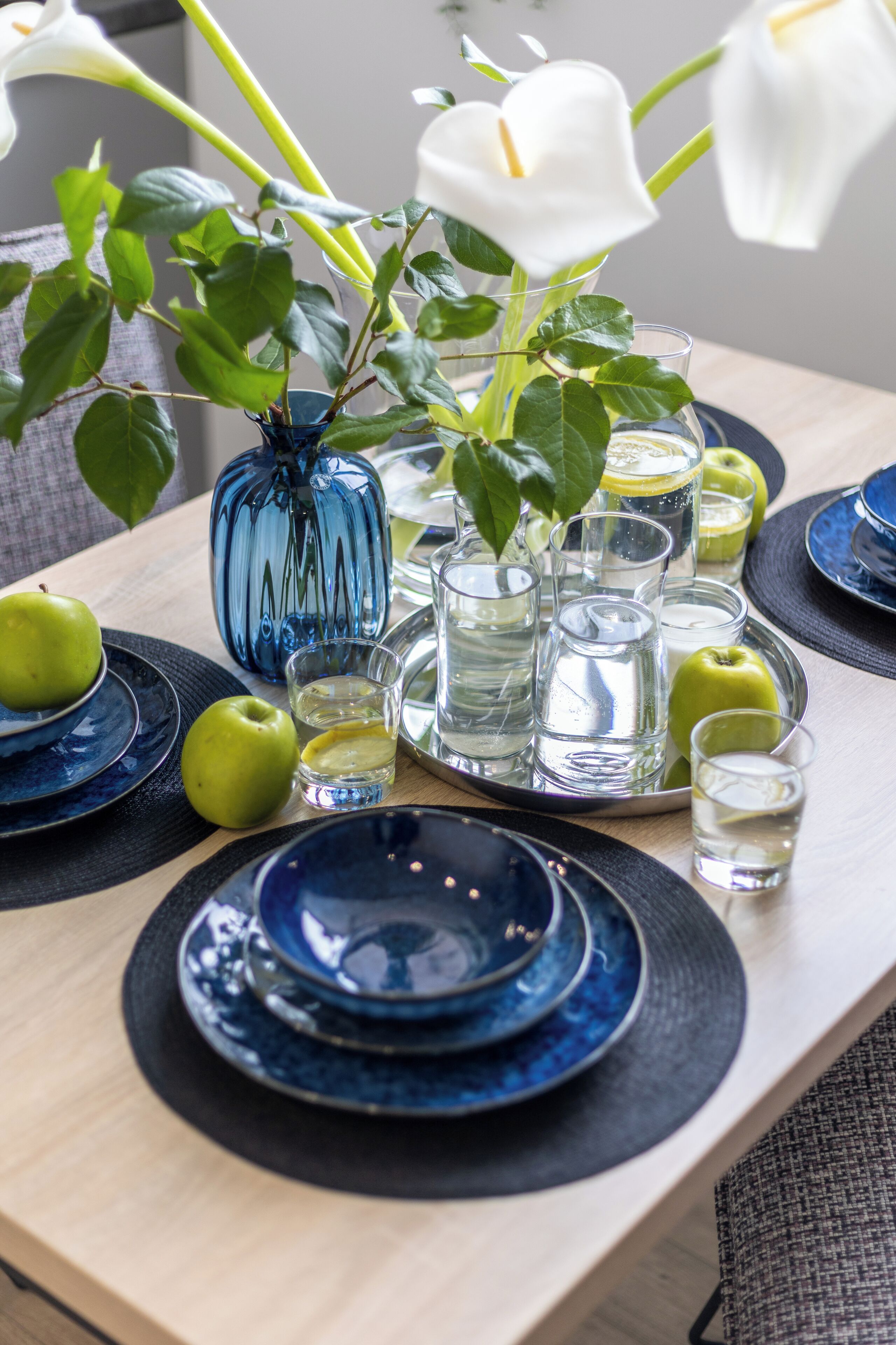 Una mesa dispuesta con platos azules, manzanas verdes, un florero azul con flores y vasos transparentes.