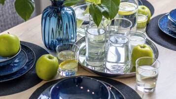Una mesa dispuesta con platos azules, manzanas verdes, un florero azul con flores y vasos transparentes.